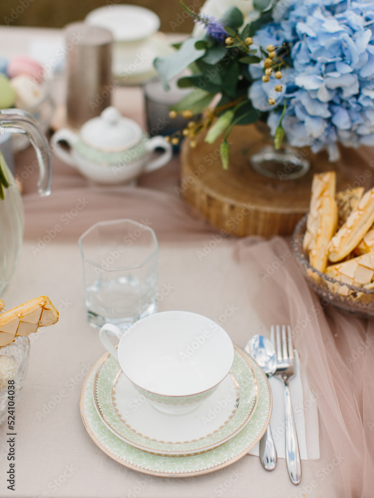 Table setting for tea. Porcelain cup on a saucer with a green border, cutlery, cookies in a crystal vase, and a bouquet of blue hydrangeas.