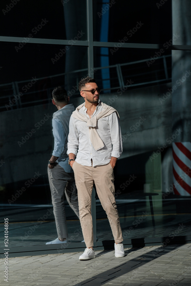 Fototapeta premium Brutal bearded man hipster in fashionable summer white clothes in sunglasses in stylish white sneakers stands on a sunny day near a glass building. Nice guy. Trendy menswear. Street style