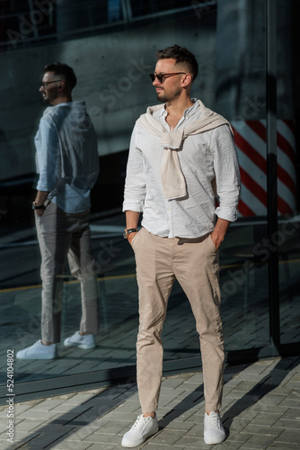 Brutal bearded man hipster in fashionable summer white clothes in sunglasses in stylish white sneakers stands on a sunny day near a glass building. Nice guy. Trendy menswear. Street style