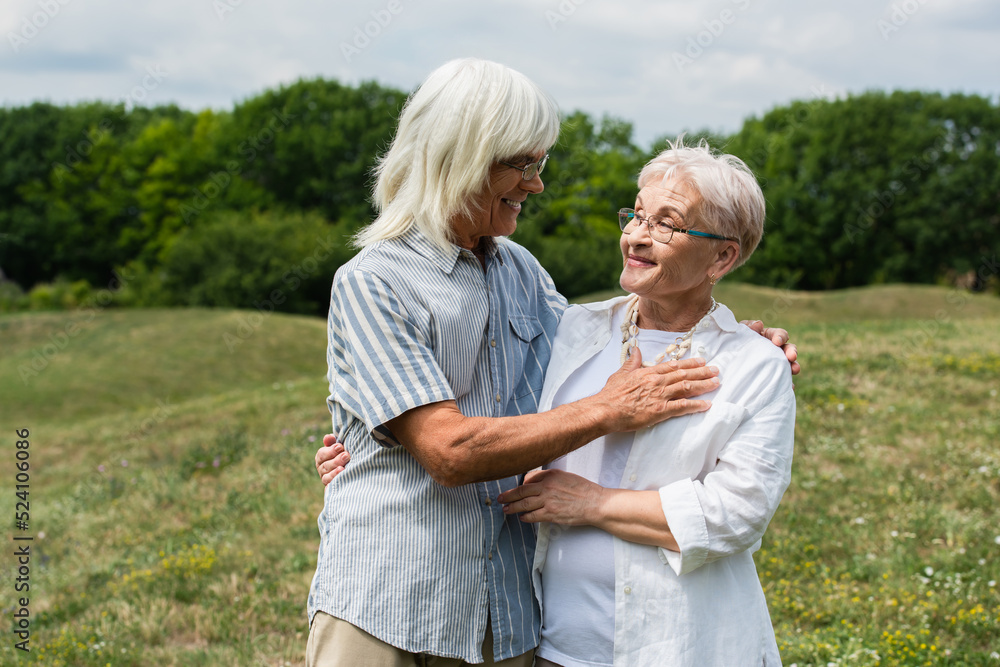 smiling senior couple in glasses hugging and looking at each other on green hill in summer. smiling senior couple in glasses hugging and looking at each other on green hill in summer.