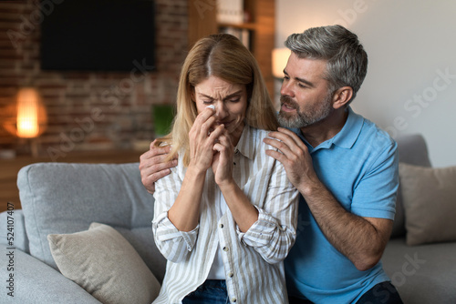 Sad middle aged caucasian man with beard hug, calm crying unhappy wife in living room interior