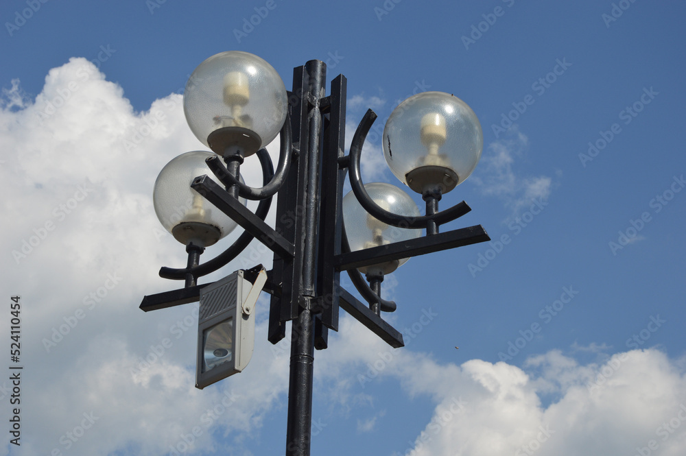 Black metal round street electric lamps with glass shades on a blue sky and clouds. Street city lighting. Black metal round street electric lamps with glass shades on a blue sky and clouds. Street city lighting.