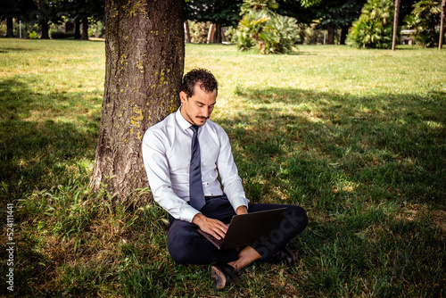 The beautiful businessman is working in a park. The young businessman is sitting on the lawn while he uses the computer.