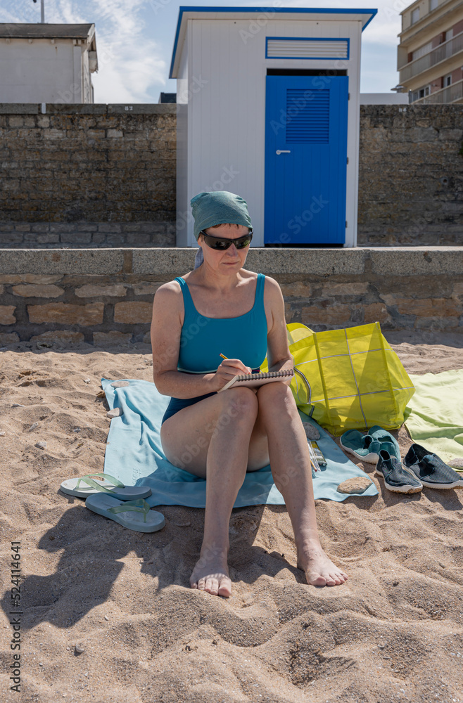 Langrune Sur Mer, France - 08 04 2022: A woman sitting on the sand of the beach wearing sunglasses, a blue cap and a blue swimsuit is drawing