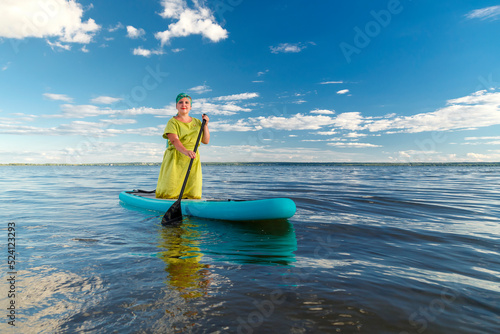 A woman in a dress and a headdress standing on a SUP board with an oar floats on the water against the blue sky. © finist_4