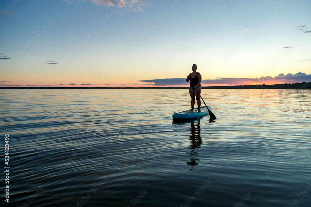 a woman in a closed swimsuit with a mohawk standing on a SUP board with an oar floats on the water at dusk.