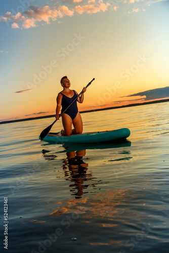 a Jewish feminist woman in a closed swimsuit with a mohawk on a SUP board with an oar floats on the water against the background of the sunset sky.
