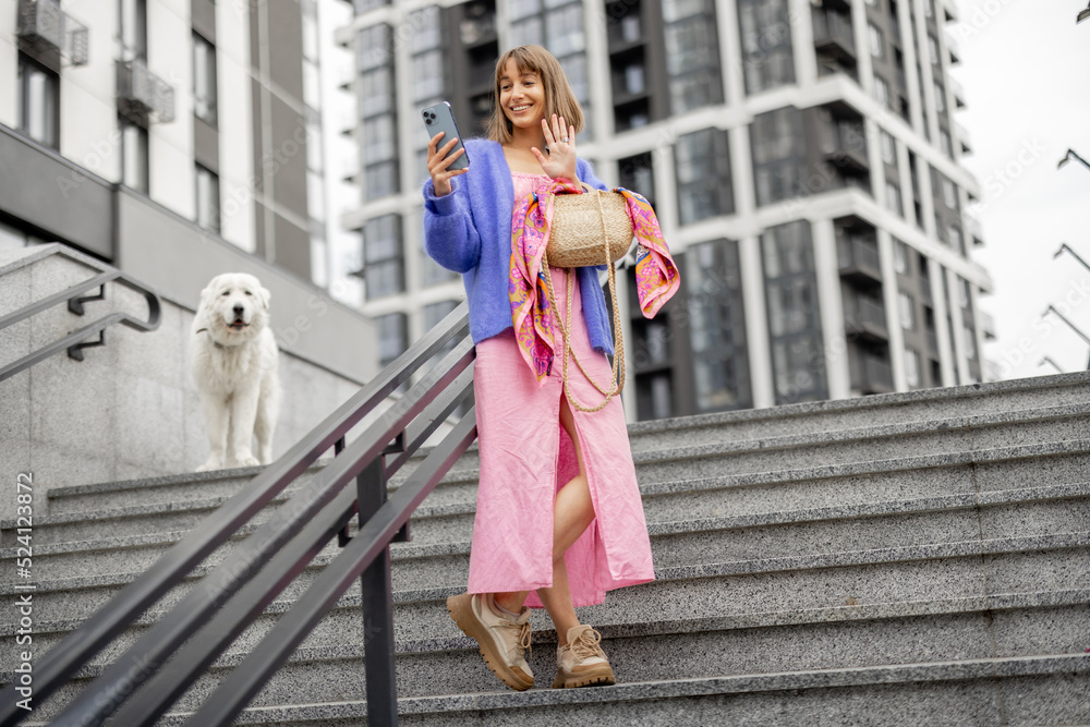 Young stylish woman dressed brightly stands with a smart phone on staircase at modern housing estate outdoors. Concept of using gadgets at everyday life