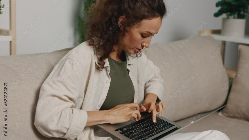 A woman with curly freelance hair sits at home on the couch, cheerfully typing on the keyboard with a smile A woman with curly freelance hair sits at home on the couch, cheerfully typing on the keyboard with a smile