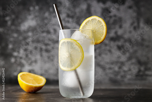 Fototapeta Refreshing homemade lemonade made of lemon slices, sparkling water served in glass with metal straw on  against gray wall