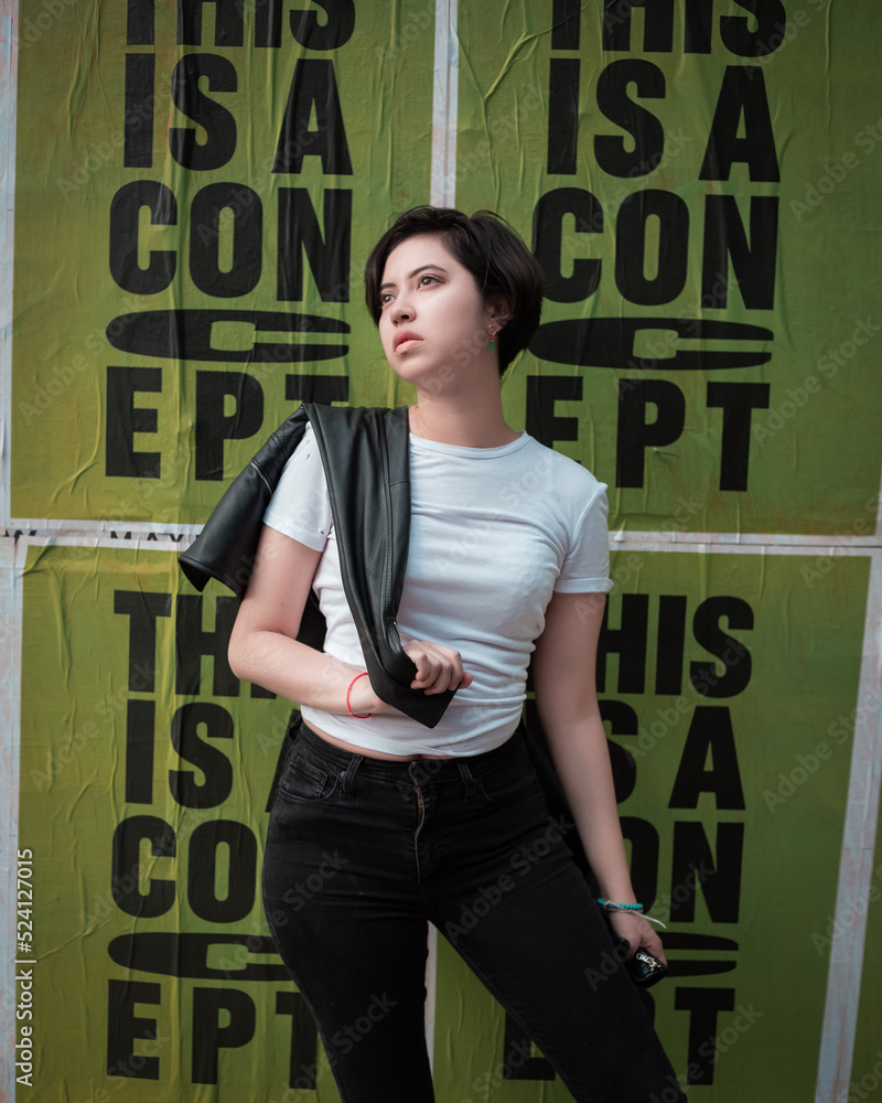 young greaser girl with short hair, posing in a fifties rockabilly style  dressed in a white t-shirt black pants leather jacket and sunglasses with a  green poster expressing fashion rebelliousness Photos