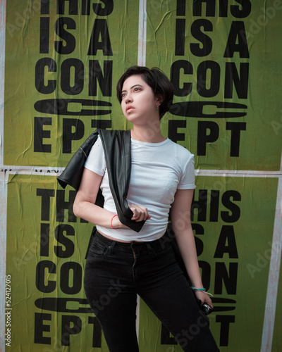 young greaser girl with short hair, posing  in a fifties rockabilly style dressed in a white t-shirt black pants leather jacket and sunglasses with a green poster expressing fashion rebelliousness  photo