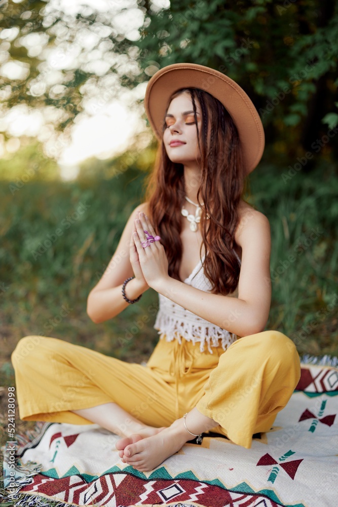 Woman in ecological clothing in a hippie look sits on a colored plaid meditating in the lotus position smiling and looking at the autumn sunset in the park
