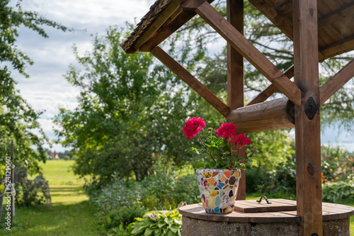 Homemade mosaic flower pot with porcelain pieces. Recycling, broken tableware, ceramics, crafts, mosaics. The vase stands on top of the well. Red geranium blooms.