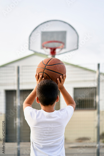 A cute young boy plays basketball on the street playground in summer. Teenager in a white t-shirt with orange basketball ball outside. Hobby, active lifestyle, sports activity for kids