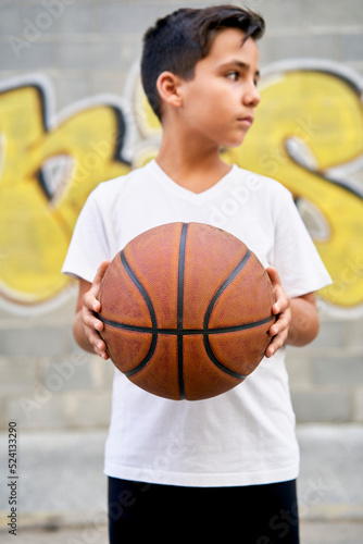 A cute young boy plays basketball on the street playground in summer. Teenager in a white t-shirt with orange basketball ball outside. Hobby, active lifestyle, sports activity for kids © photostocklight