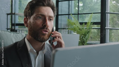 Angry bearded Caucasian adult 40s mad frustrated businessman arguing yelling conflict quarrel talking on phone looking at laptop computer sitting at office dissatisfied male client problem bad service photo