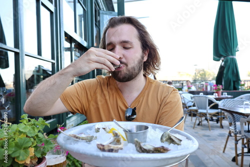 young white male with long hair and beard, wearing mustard t-short, sitting outside in the restaurant eating fresh oysters with his hands