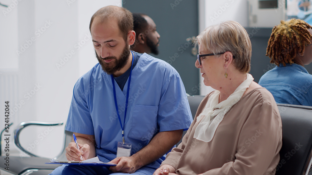 Obraz premium Male nurse consulting senior patient in waiting room at medical facility, doing checkup examination to cure disease. Woman with diagnosis talking about medicine with specialist in reception lobby.