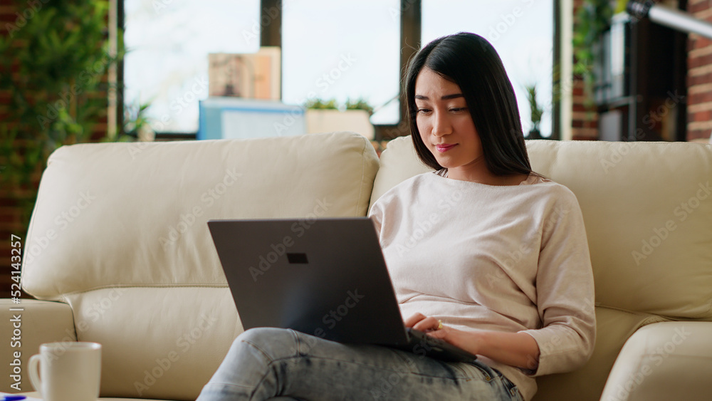 Naklejka premium Smiling office worker at home working remotely on laptop while sitting on sofa in living room. Young adult person doing remote work on modern portable computer while at home.