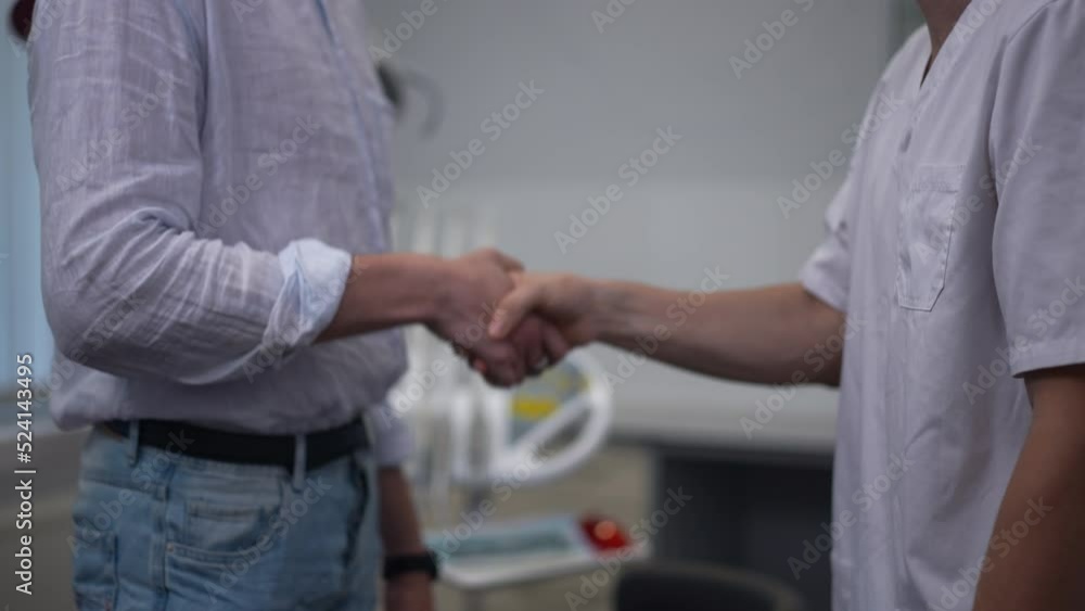 Side view unrecognizable dentist and patient handshaking in slow motion standing indoors. Caucasian man shaking hands with doctor in medical clinic. Medicine and service concept Side view unrecognizable dentist and patient handshaking in slow motion standing indoors. Caucasian man shaking hands with doctor in medical clinic. Medicine and service concept