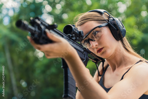 Young caucasian woman wearing protective goggles and headphones aiming submachine gun on training ground. Blurred background horizontal shot. High quality photo © PoppyPix
