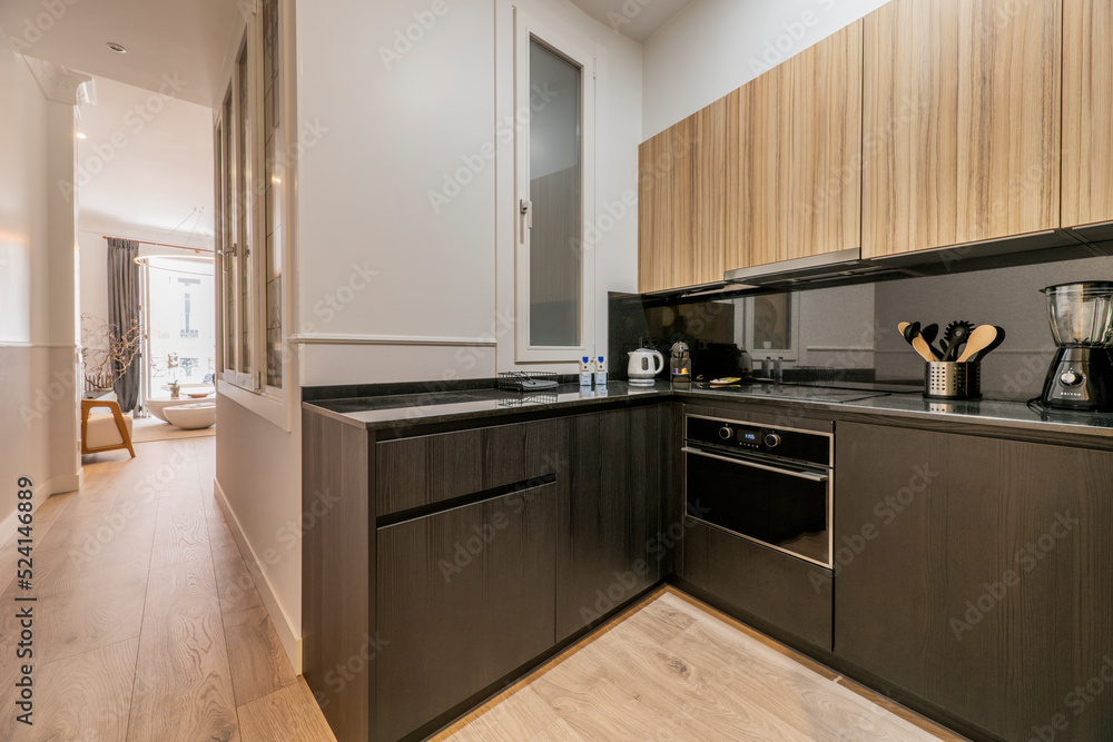 Fototapeta premium Open kitchen with low black and high wooden cabinets, integrated appliances and a corridor leading to a living room with a balcony