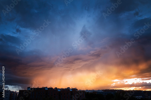 фотографія a summer storm at sunset over a city with a sky in shades of orange and blue, co