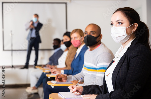 Young focused woman in protective face mask sitting and listening to speaker at business conference