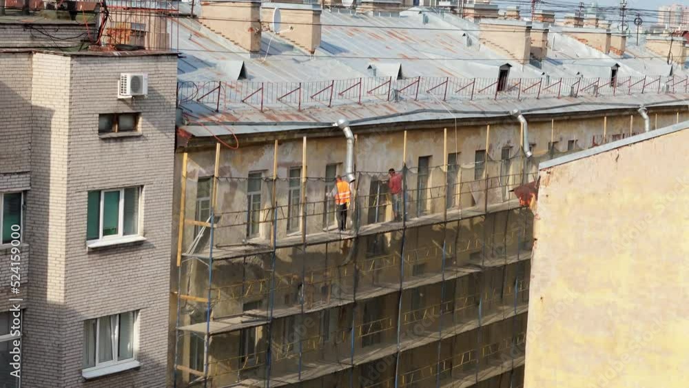 Workers in reflective vests restore old multi-story facade building. Men stand on construction scaffolding working near windows on sunny day Workers in reflective vests restore old multi-story facade building. Men stand on construction scaffolding working near windows on sunny day
