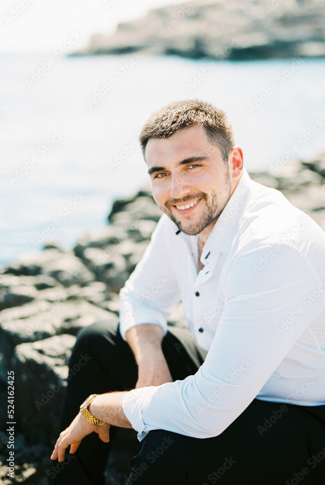 Smiling man in a white shirt sits on a stone by the sea