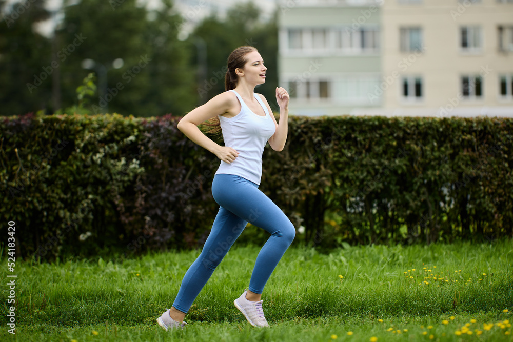 Woman 30s jogging in summer park near residential area with apartment buildings.