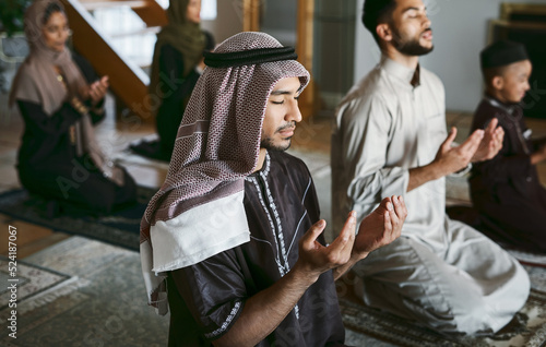 Young muslim family pray together, sit on knees at home practicing Islam. Religious women wearing hijab and spiritual men wear traditional attire, close eyes and concentrate on prayer with hands up.