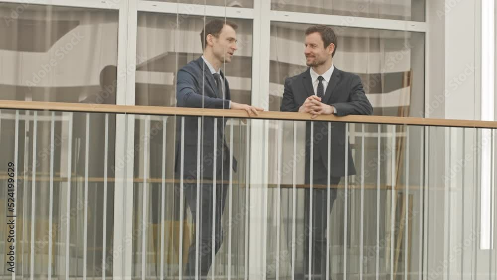 Full length slowmo of two Caucasian businessmen in elegant formalwear standing by glass railing at modern business center having conversation Full length slowmo of two Caucasian businessmen in elegant formalwear standing by glass railing at modern business center having conversation