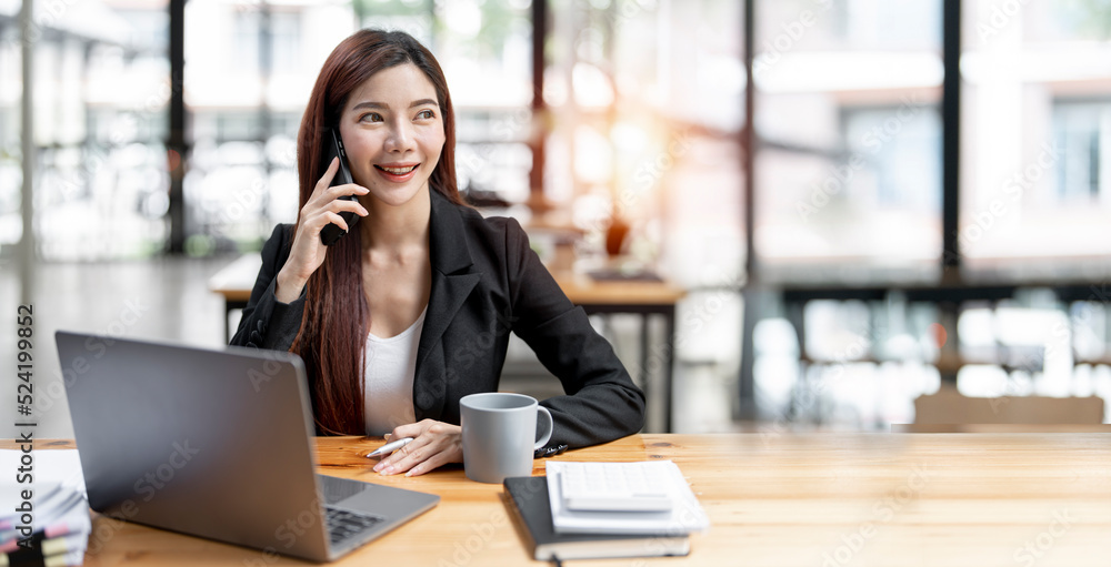 Fototapeta premium Happy businesswoman sitting at desk behind her laptop and talking with somebody on her mobile phone while working at office.