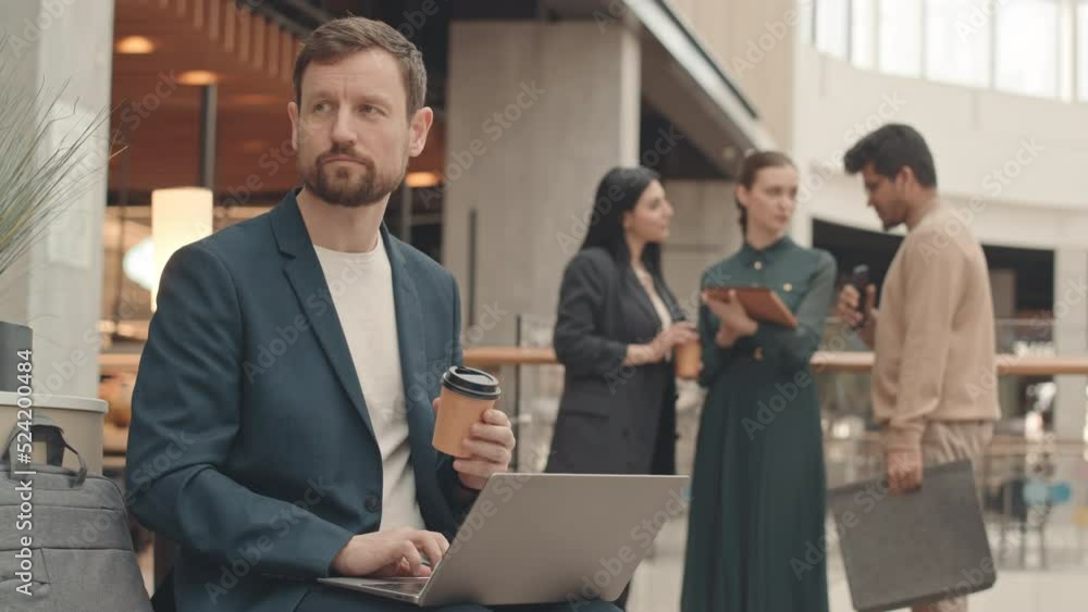 Medium slowmo of Caucasian man in smart casualwear holding takeaway coffee cup in hand and working on laptop sitting on bench at modern business center. His colleagues chatting in blurred background Medium slowmo of Caucasian man in smart casualwear holding takeaway coffee cup in hand and working on laptop sitting on bench at modern business center. His colleagues chatting in blurred background