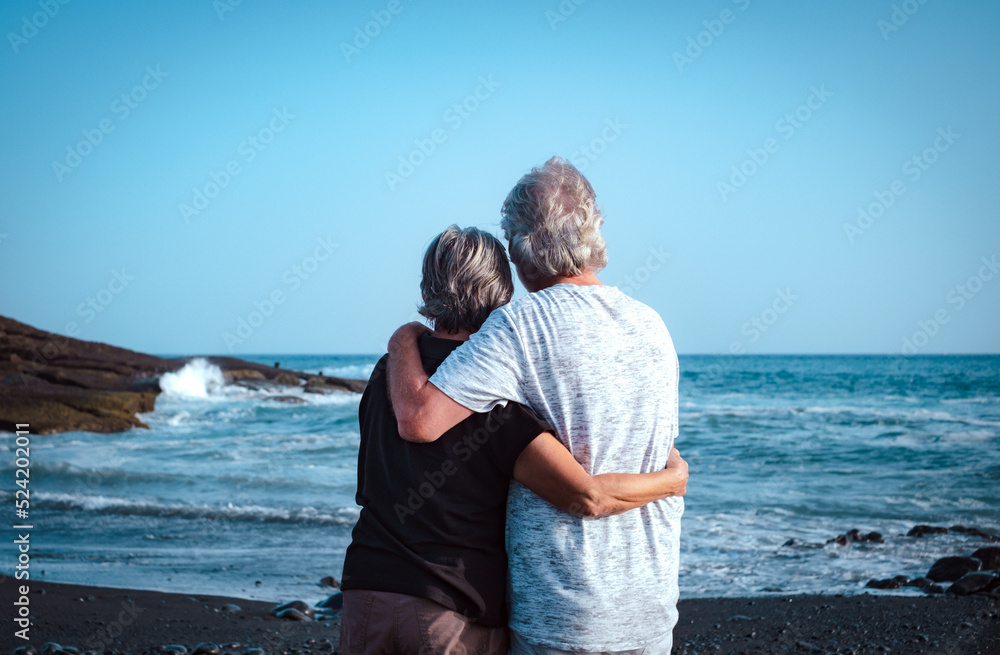 Back view of enbraced caucasian senior couple standing on the beach at sunset looking at the horizon- active pensioners enjoying free time, sea vacation or retirement Back view of enbraced caucasian senior couple standing on the beach at sunset looking at the horizon- active pensioners enjoying free time, sea vacation or retirement