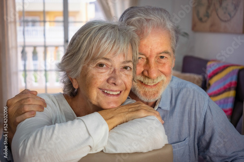 Portrait of beautiful caucasian senior couple sitting at home hugging and looking at camera