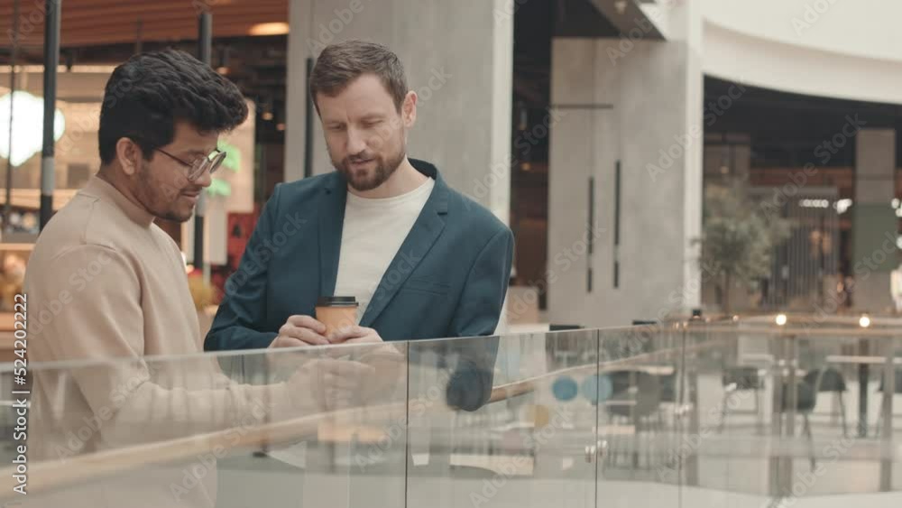 Medium slowmo of two diverse male colleagues talking during coffee break, standing by glass railing at contemporary business center Medium slowmo of two diverse male colleagues talking during coffee break, standing by glass railing at contemporary business center