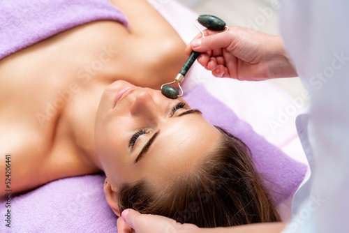 Facial massage. Side view european woman getting massage with jade face roller gouache in spa salon