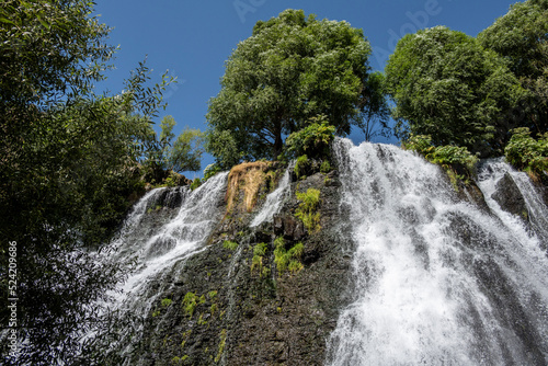 a stormy waterfall against the backdrop of green trees and mountains and a blue sky in the mountains of Armenia