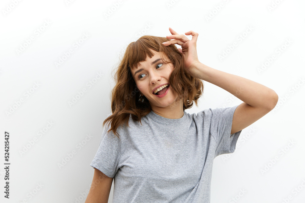 a happy, joyful woman in a gray cotton T-shirt stands against a light background, smiling happily and looking away, holding her hair with her hand. a happy, joyful woman in a gray cotton T-shirt stands against a light background, smiling happily and looking away, holding her hair with her hand.