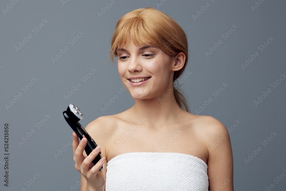 a woman with perfect skin stands on a gray background wrapped in a towel and examines an electric massager in her hands, smiling pleasantly. Close horizontal photo a woman with perfect skin stands on a gray background wrapped in a towel and examines an electric massager in her hands, smiling pleasantly. Close horizontal photo