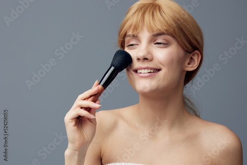 a happy, joyful woman stands on a gray background and paints her face with a fluffy makeup brush, closing her eyes with pleasure