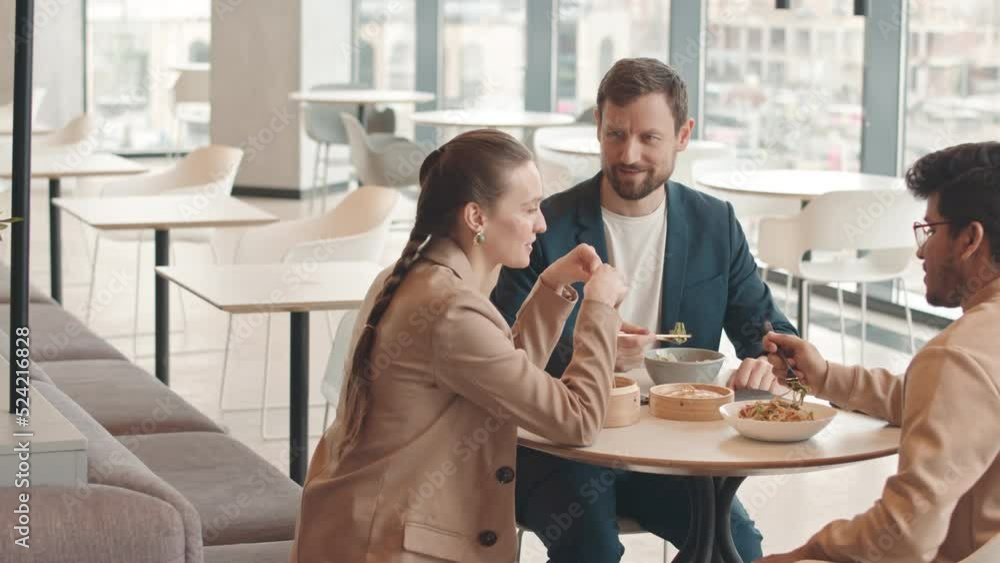 Medium slowmo of three coworkers having lunch together and talking sitting at food court with panoramic city view Medium slowmo of three coworkers having lunch together and talking sitting at food court with panoramic city view