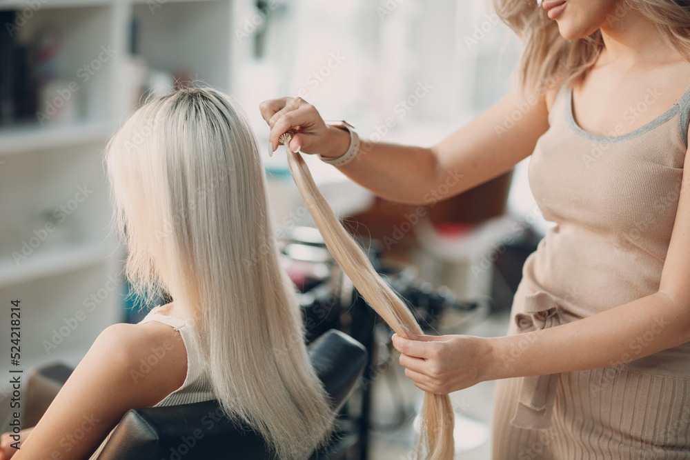 Hairdresser female making hair extensions to young woman with blonde hair in beauty salon. Professional hair extension