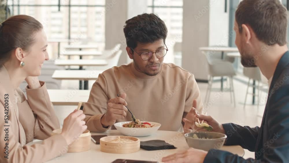 Waist up slowmo of multiethnic colleagues talking while having lunch with Pan Asian food at food court with panoramic windows