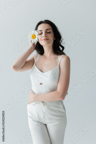 Pleased woman in white clothes holding chamomile isolated on grey. © LIGHTFIELD STUDIOS