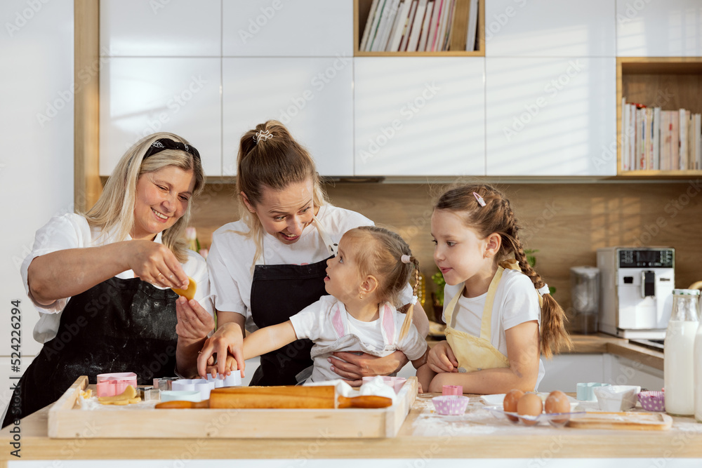 Grandmother showing cookies to grandchildrn. Surprised young mother holding in arm preschooler daughter talking and smiling. Older daughter watching process. Family having fun together. Grandmother showing cookies to grandchildrn. Surprised young mother holding in arm preschooler daughter talking and smiling. Older daughter watching process. Family having fun together.