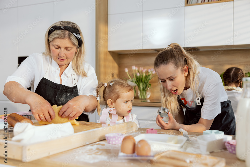 Granny preparing dough for homemade domestic cookies using eggs milk flour. Young mother showing cookies cutter preschooler pretty daughter. In background older daughter washing diches. Granny preparing dough for homemade domestic cookies using eggs milk flour. Young mother showing cookies cutter preschooler pretty daughter. In background older daughter washing diches.